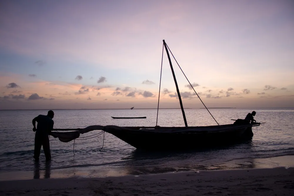 Fishermen beside a wooden dhow on the shore at dusk, Zanzibar